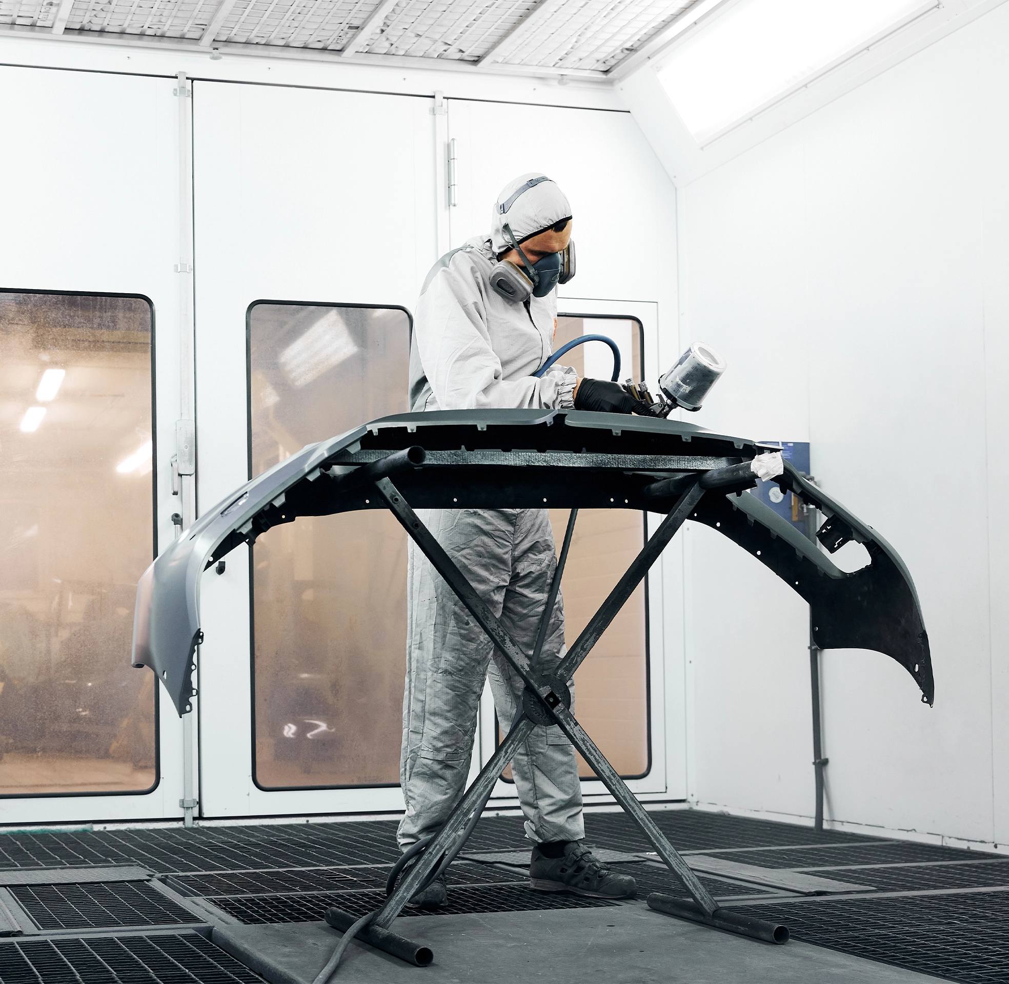 Auto body technician wearing protective gear while spray-painting a detached bumper inside a controlled paint booth.