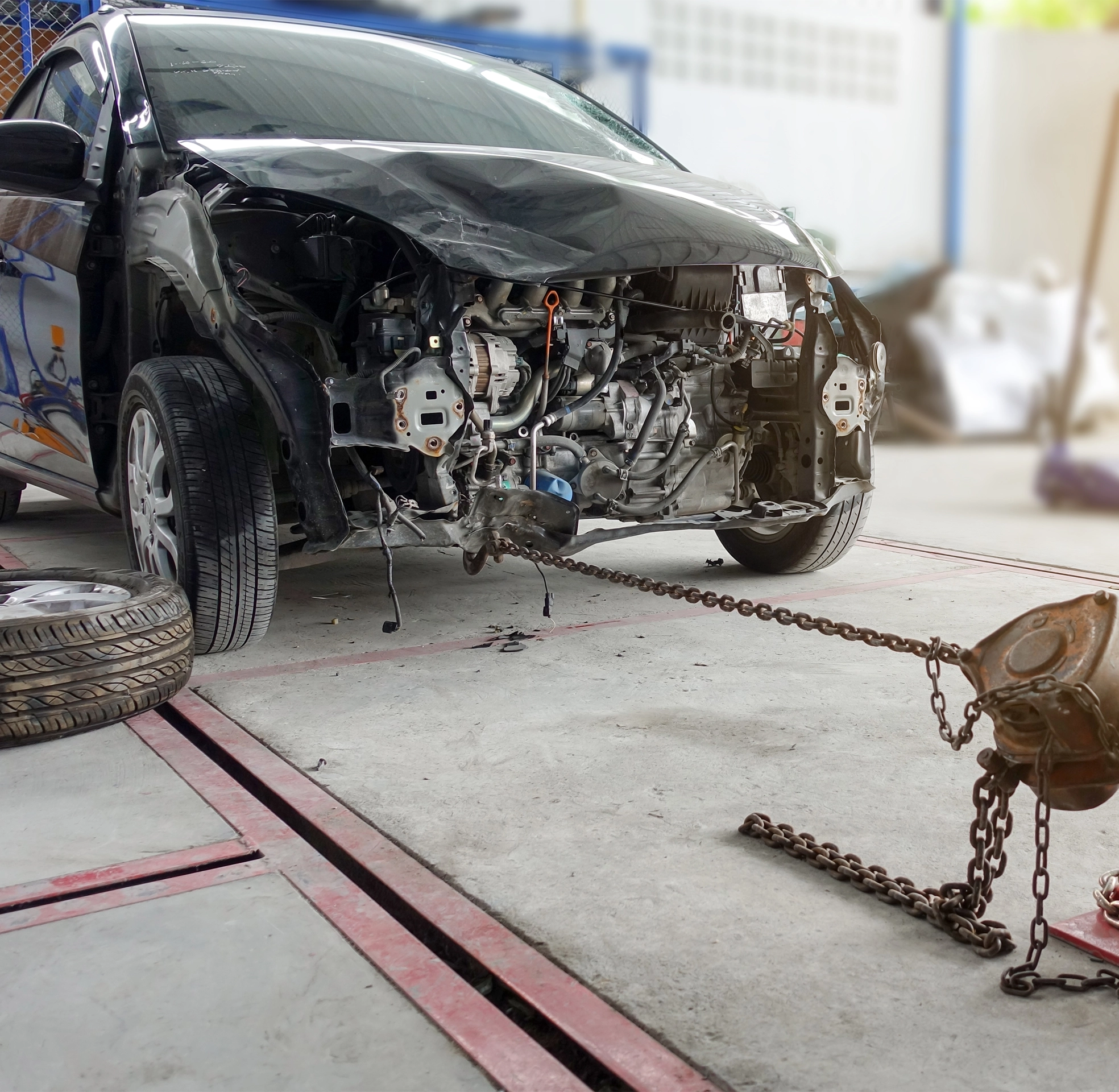 A severely damaged car in a repair shop with the entire front end removed, showing exposed engine components while a heavy-duty chain and puller are used for frame straightening.