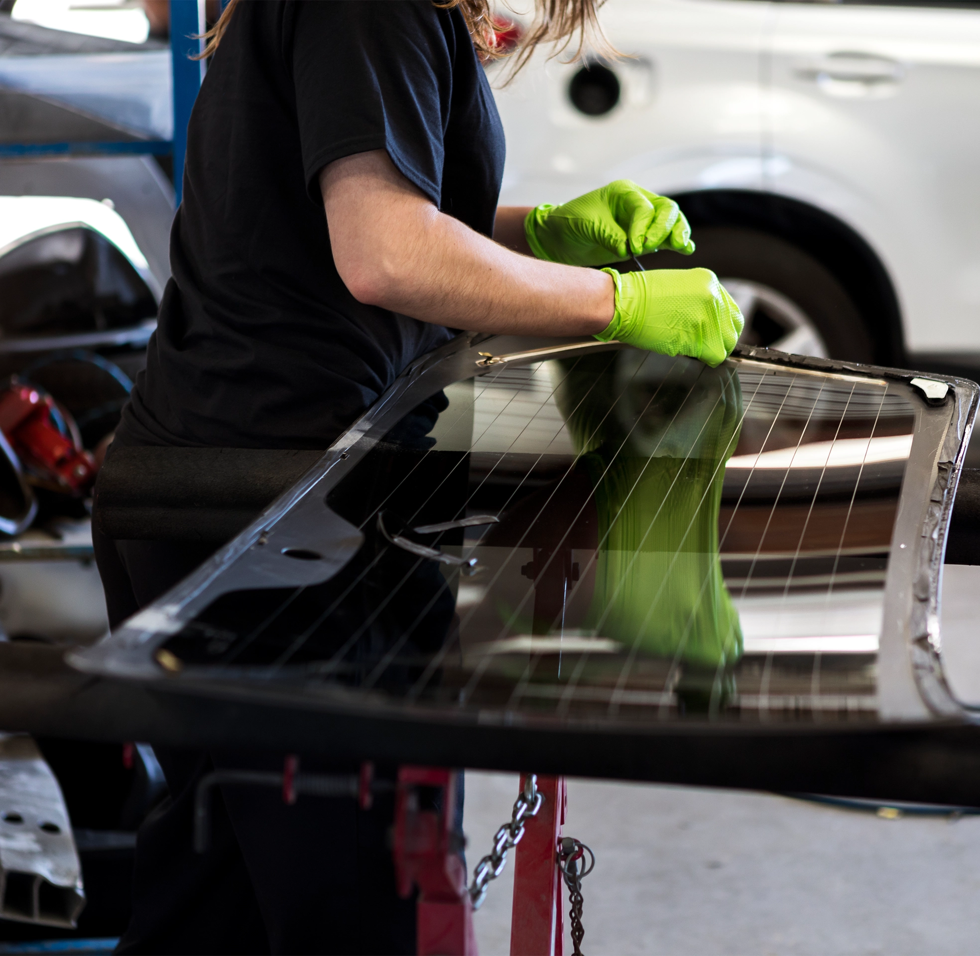Auto body technician wearing bright green gloves repairing or preparing a vehicle panel, carefully working on a glossy black surface inside a collision repair shop.