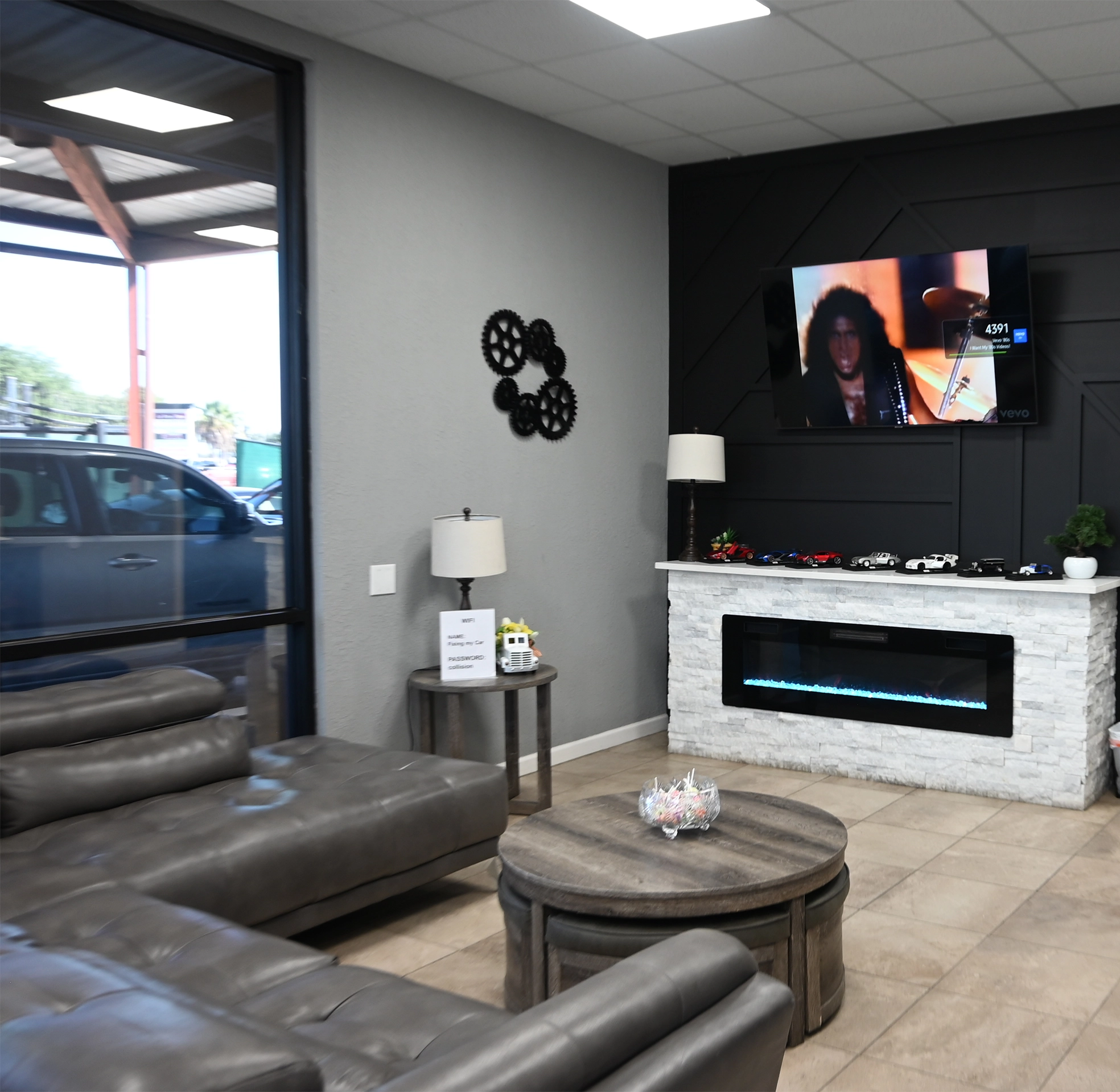 Comfortable auto shop waiting area featuring gray leather seating, a round wooden coffee table, wall decor with gear designs, a modern electric fireplace, and a TV mounted above displaying a music video. Small model cars and decor items sit on the mantle.