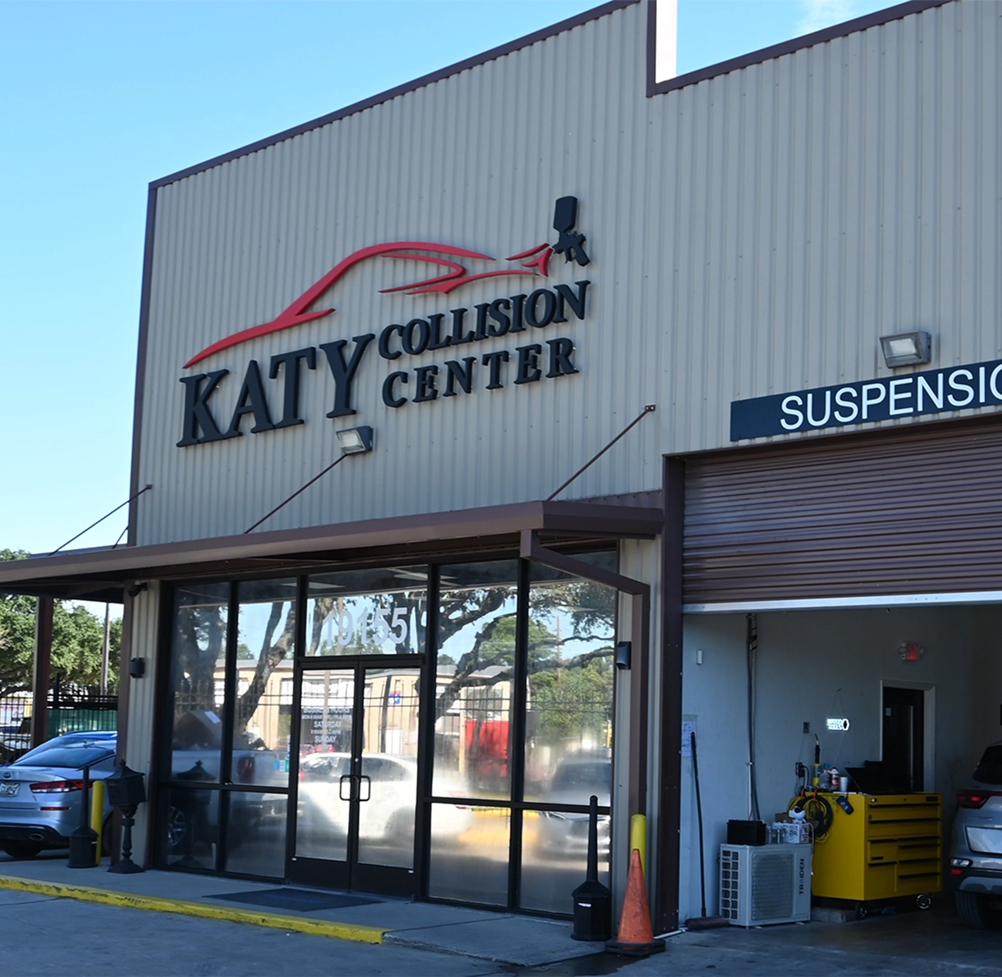 Exterior of Katy Collision Center showing the front entrance with large glass doors, the shop’s logo featuring a red car outline, and an open service bay labeled “Suspension” with equipment visible inside.