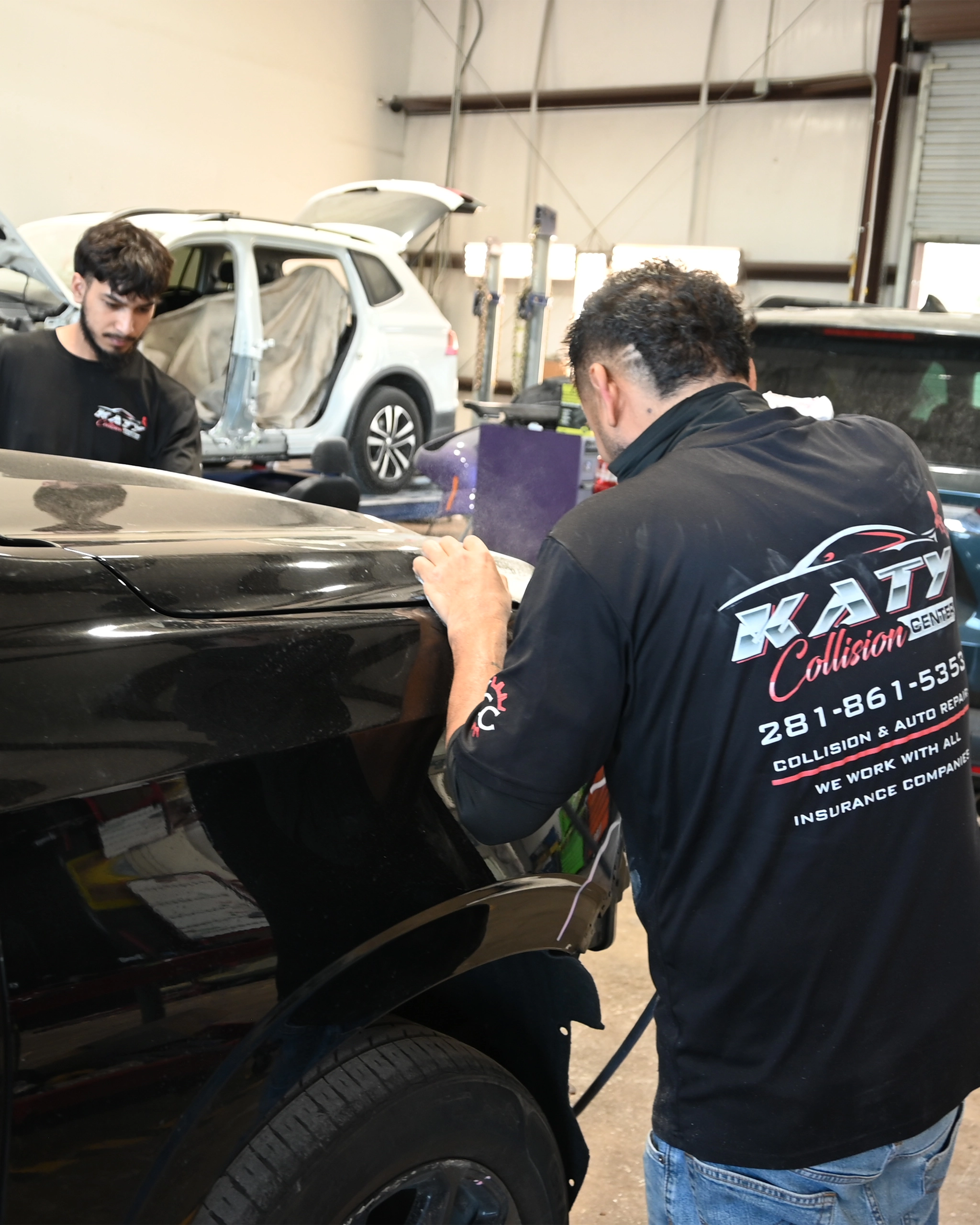 Technicians at Katy Collision Center repairing a black vehicle inside the shop, with one worker sanding a panel and another working in the background near a white SUV undergoing body repair.
